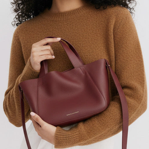 Person holding a maroon handbag with a visible brand name, wearing a brown sweater.