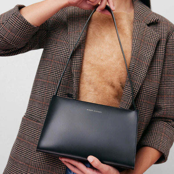 A brown leather shoulder bag with a minimal design, a sturdy zipper, and a delicate shoulder strap, displayed against a white background. - Saddle