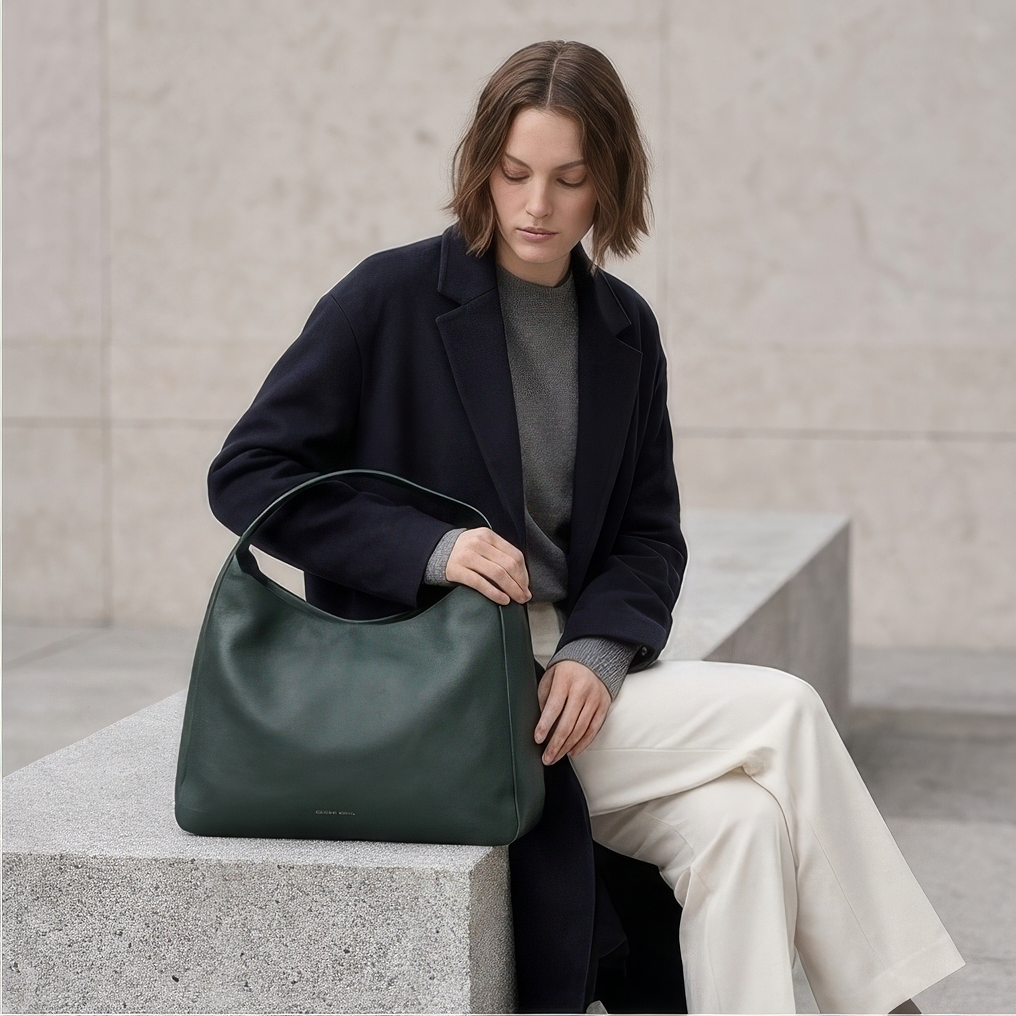 A woman sits on a stone bench wearing a dark navy coat, gray sweater, and cream trousers, holding a dark green leather shoulder bag. - Evergreen