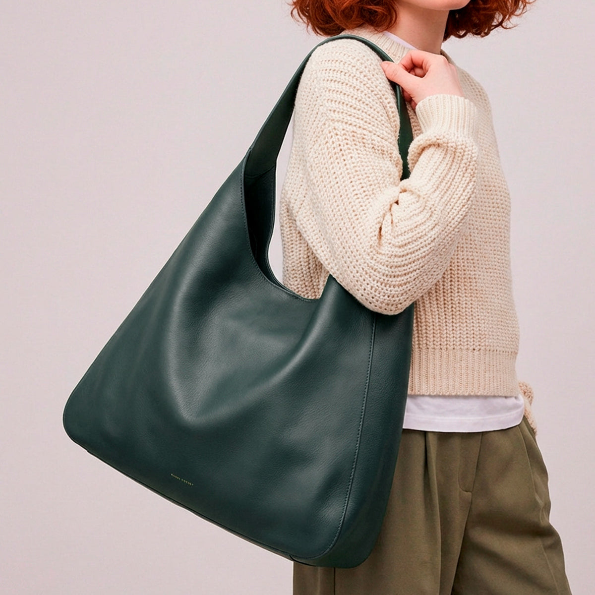 Person wearing a beige sweater and holding a green handbag against a plain background - Evergreen