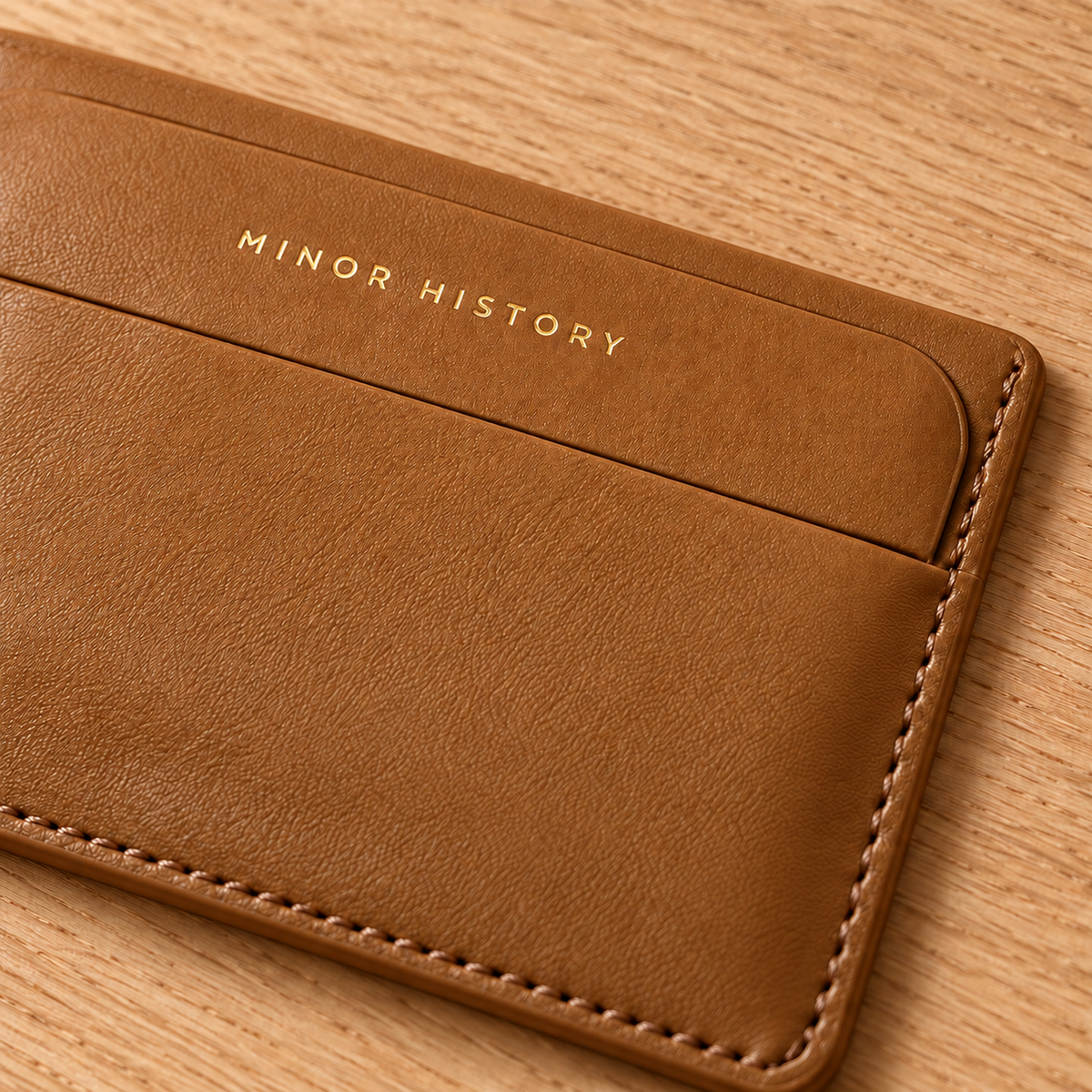 Close-up of the Midtown card holder’s saddle leather, stitching and gold Minor History logo on a wood tabletop - Saddle